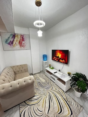 Ein modernes Wohnzimmer mit einem beigen Sofa, einem wandmontierten Flachbildfernseher, einer weißen Unterhaltungsanlage, einem gemusterten Teppich und einer Deckenleuchte.