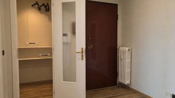 Quarto com piso de madeira, porta branca com painel de vidro jateado, radiador e armário com prateleiras.