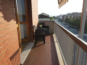 Un balcon avec un fauteuil en osier, une petite table et une vue sur une rue animée, avec des voitures et des immeubles.