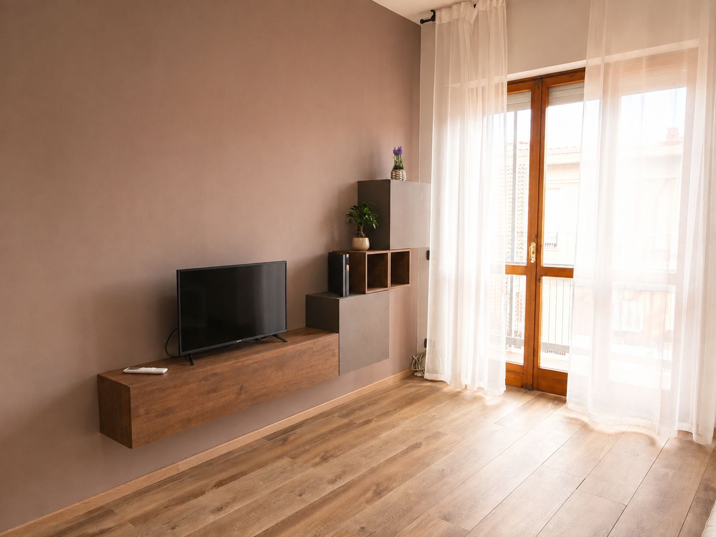 Ein modernes Wohnzimmer mit einem Flachbildfernseher auf einem Holzboden, einer Topfpflanze und einer Glastür, die zu einem Außenbereich führt.