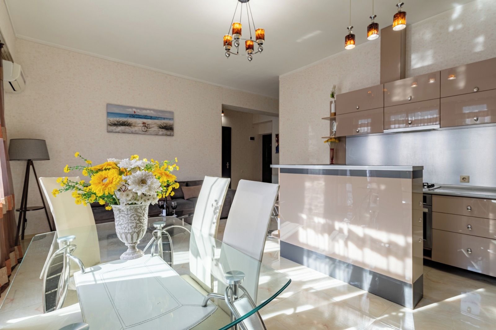 A modern kitchen with stainless steel appliances, a dining area with a glass table and chairs, and a chandelier overhead.