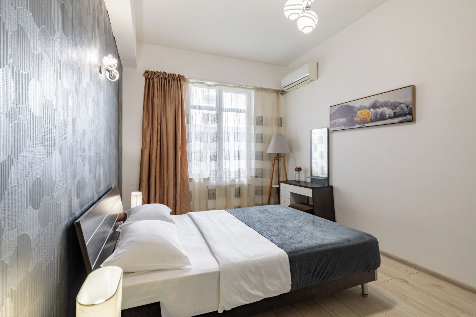 Ein Hotelzimmer mit einem Bett, einem Schreibtisch, einer Lampe, einem Fenster mit Vorhängen und einer Klimaanlage.