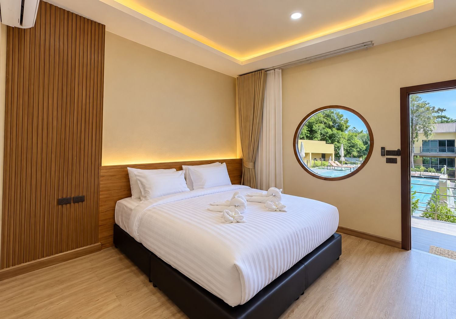 Habitación de hotel con una cama grande, pared de paneles de madera, ventana redonda y vista a una piscina y vegetación al exterior.