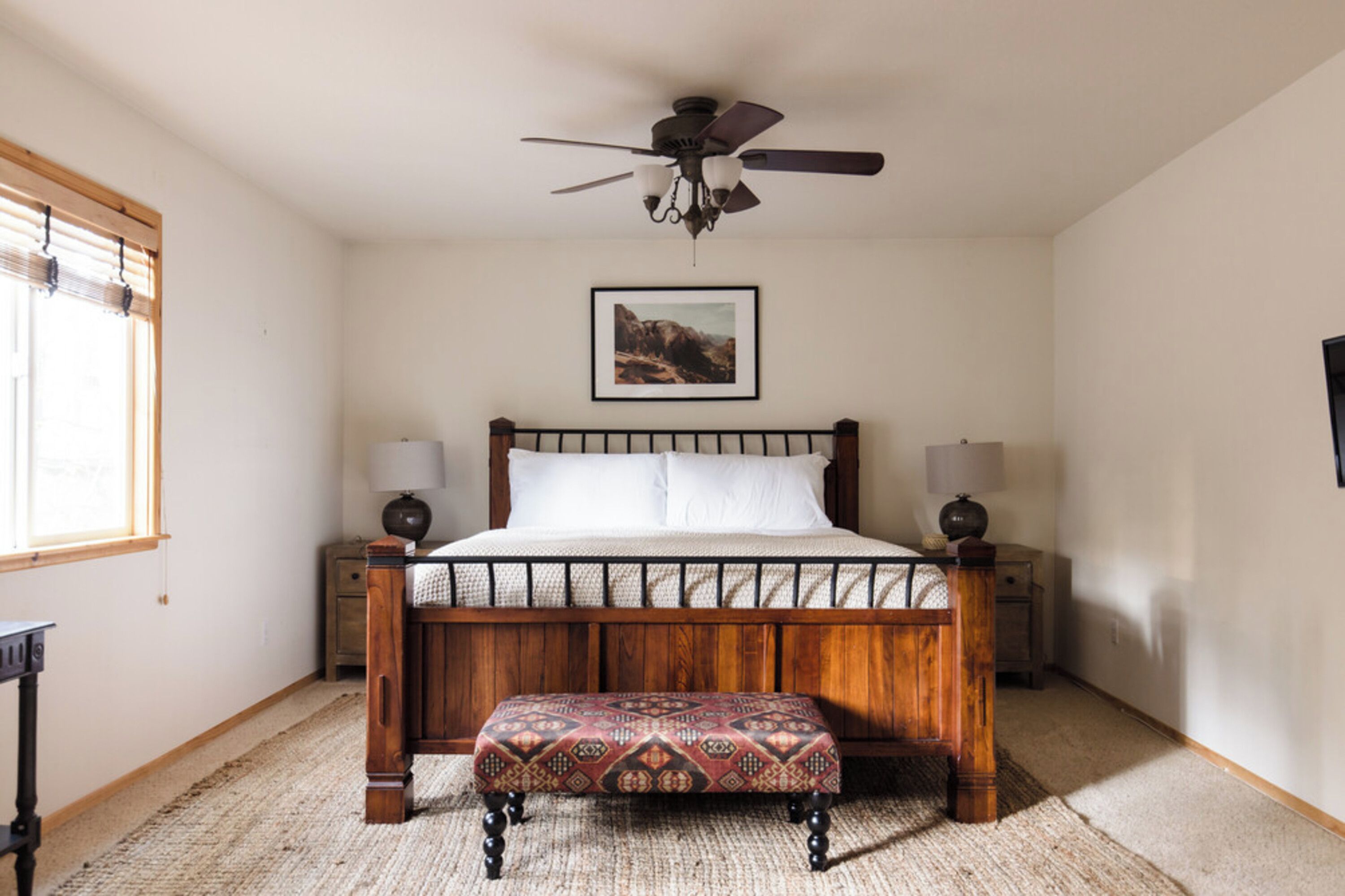 Un dormitorio con una cama de madera, un ventilador de techo, una mesita de noche, una lámpara, una ventana con persianas y un cuadro en la pared.
