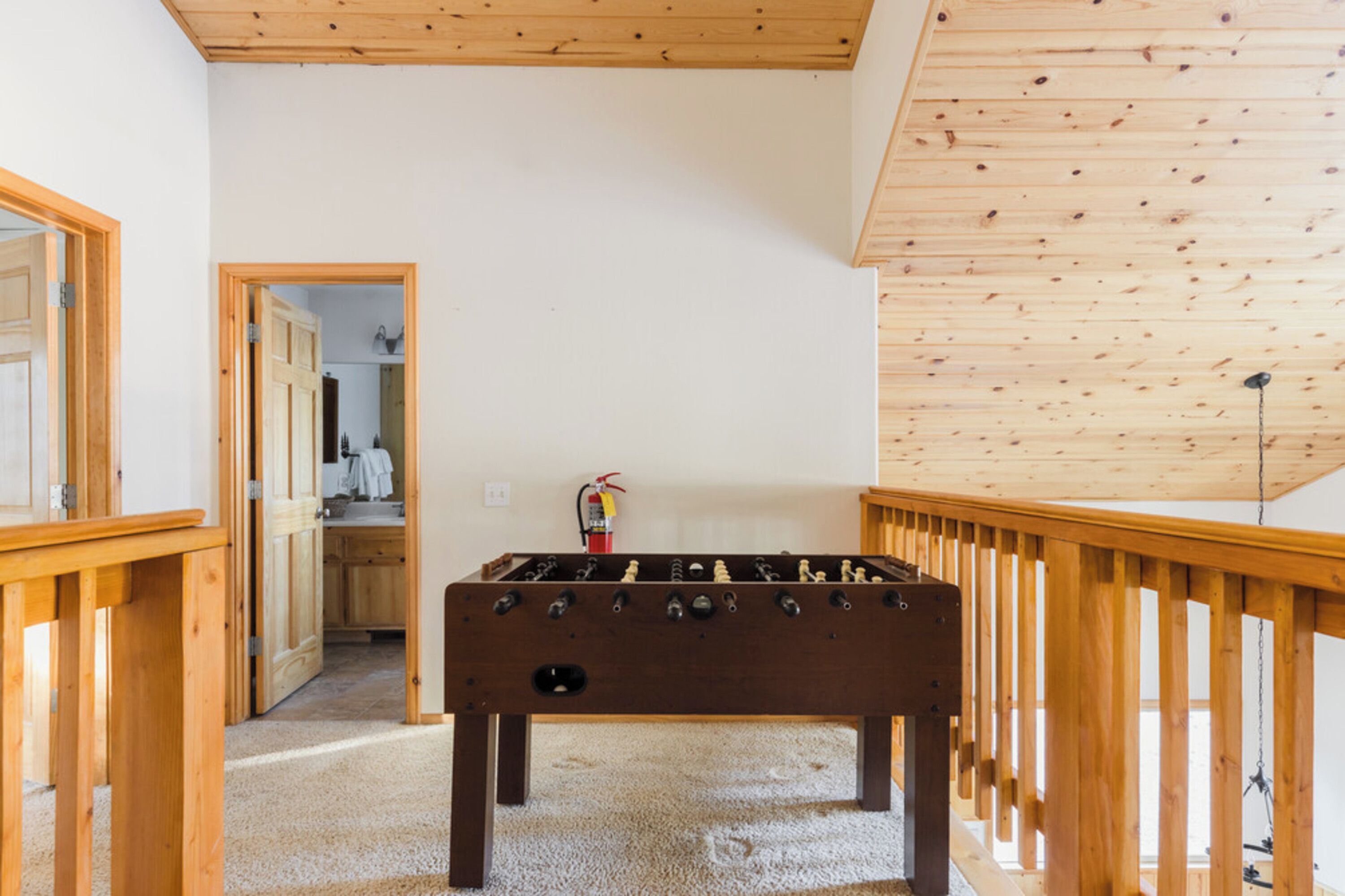 A room with a wooden foosball table, a wooden railing, and a door leading to a bathroom.