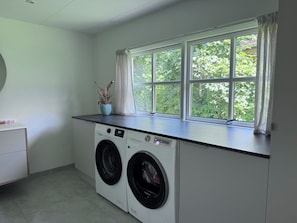 Un lavadero con dos lavadoras de carga frontal, una ventana con cortinas y una pequeña planta en el alféizar.
