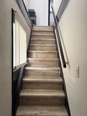 Un escalier en bois avec une rampe, menant à un palier doté d’une fenêtre et d’une étagère en métal.