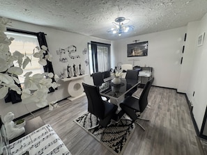 Un espace de salle à manger moderne, avec un tapis à motifs noir et blanc, un ventilateur de plafond et un téléviseur fixé au mur.