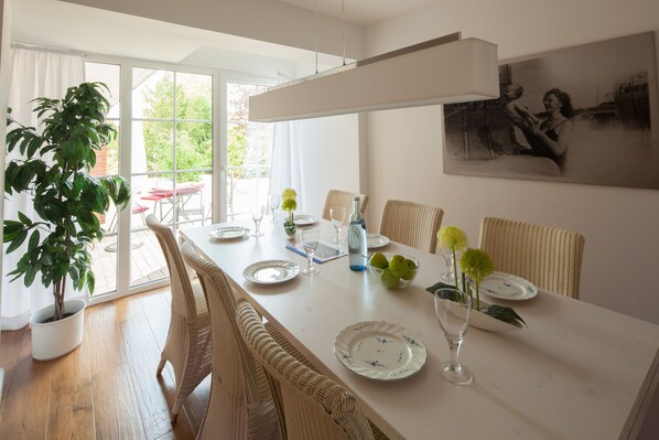 Un coin repas avec une longue table dressée pour quatre personnes, entourée de chaises en osier, d’une plante en pot et d’un tableau accroché au mur.