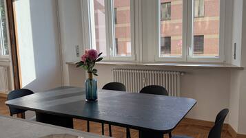 Zona pranzo con tavolo, sedie, un vaso con fiori e ampie finestre vista edifici.