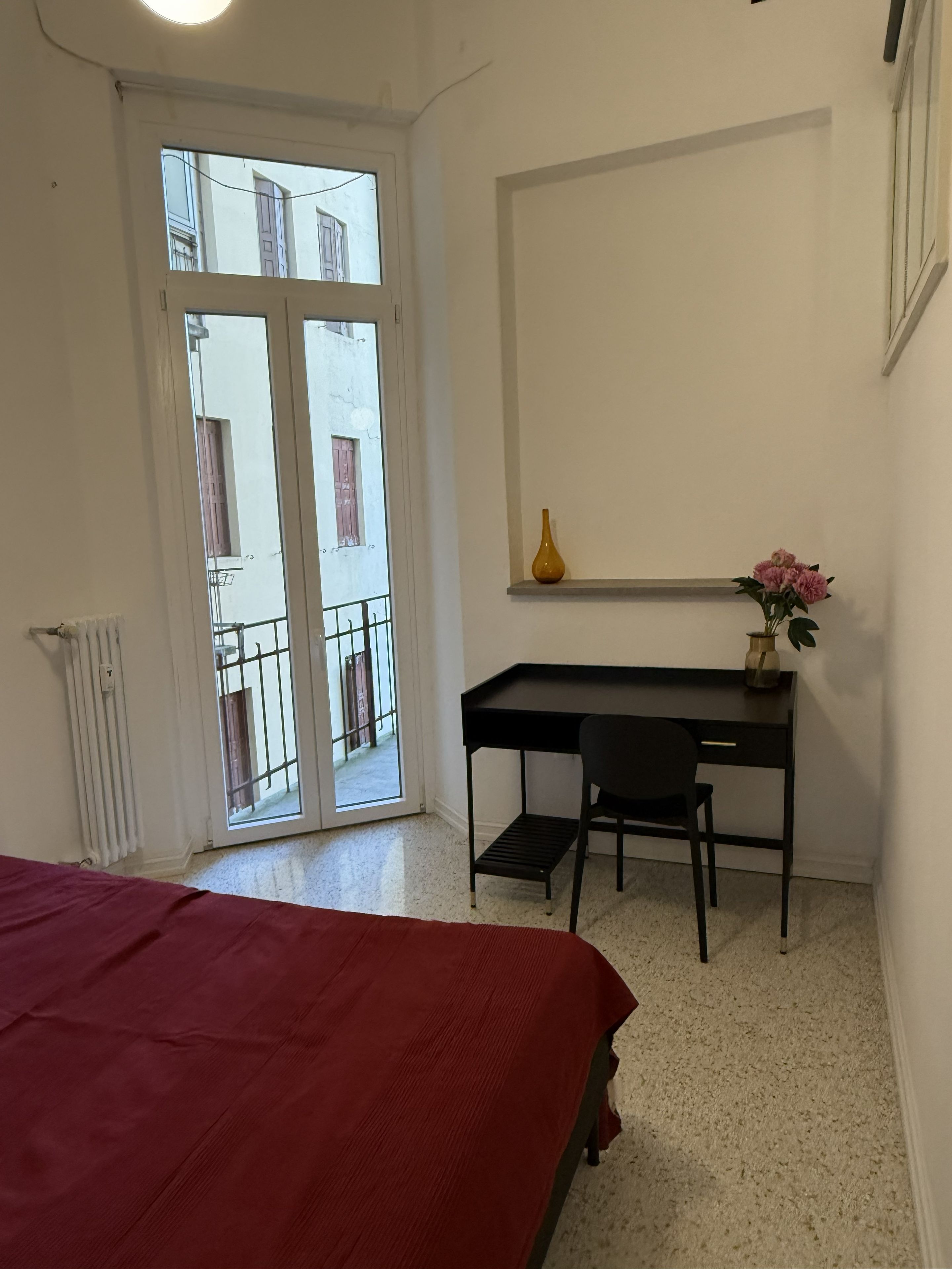 Una stanza con un letto, una scrivania, una sedia, un termosifone e una finestra vista edifici.