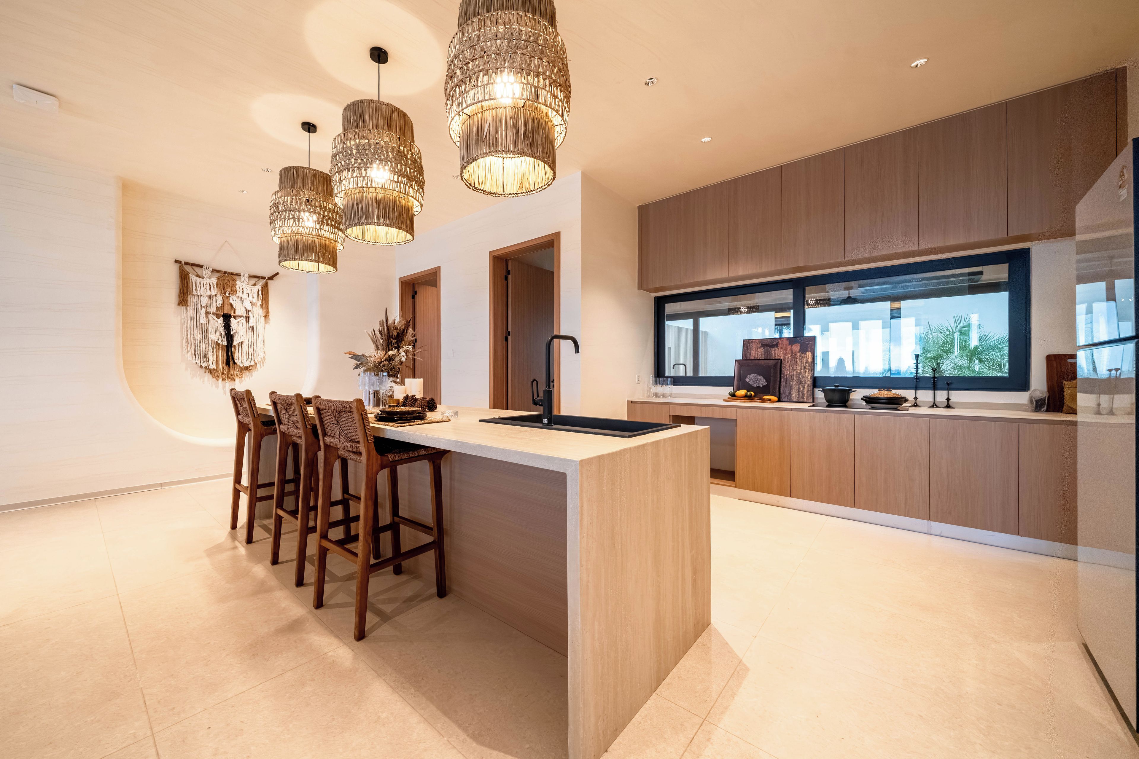 A modern kitchen with a central island, wooden bar stools, pendant lighting, and a view of the outdoors through a large window.