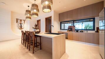 A modern kitchen with a central island, wooden bar stools, pendant lighting, and a view of the outdoors through a large window.