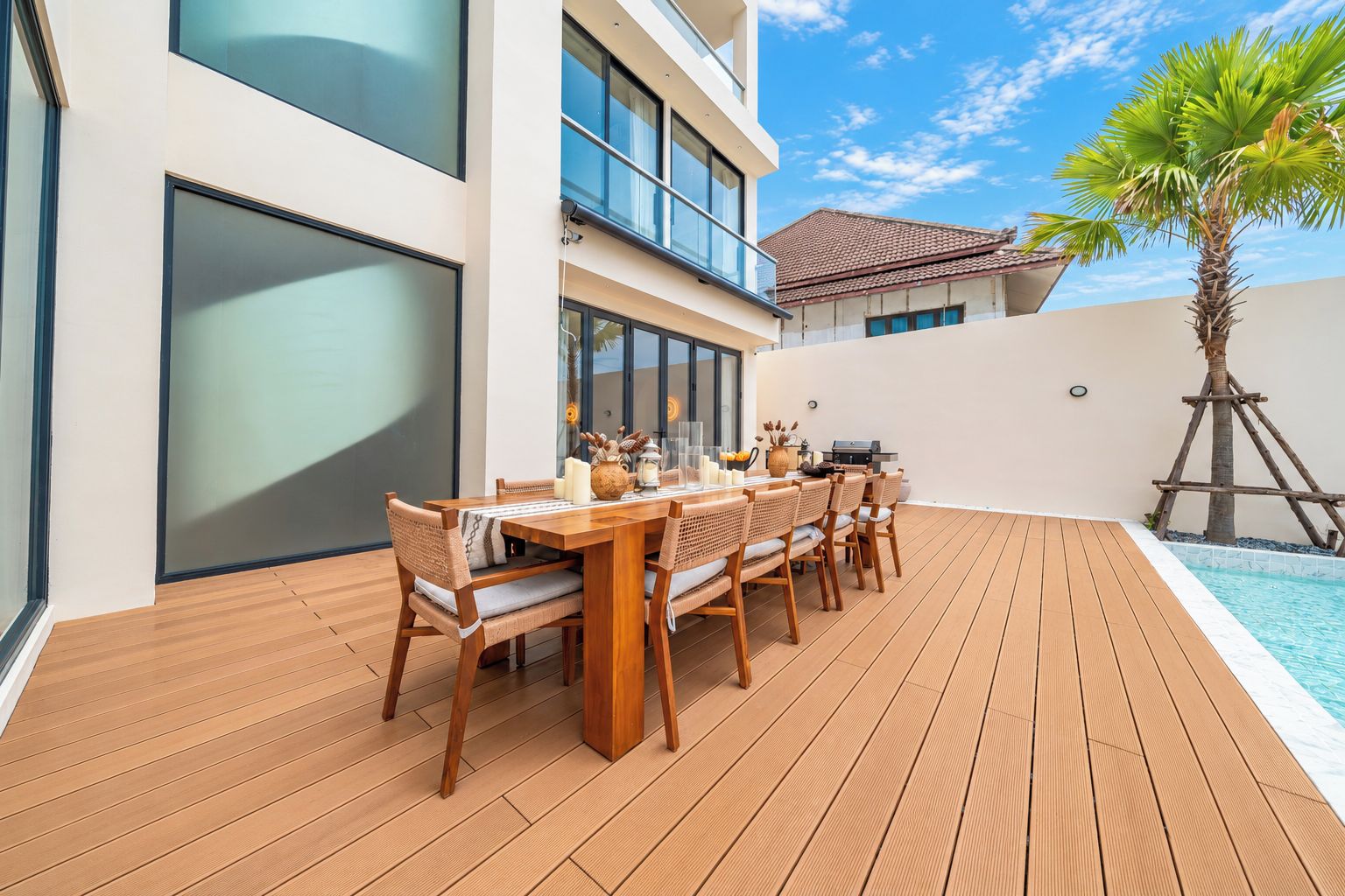A wooden deck with a dining table and chairs, a pool, and a palm tree.