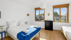 Un dormitorio con cama, cómoda y dos ventanas con vistas a edificios.
