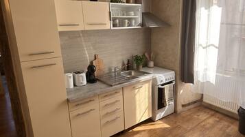 A modern kitchen with a wooden floor, stainless steel appliances, and a window with sheer curtains.