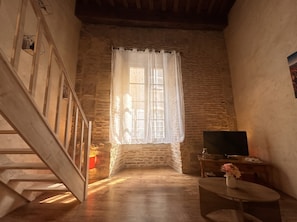 A room with a wooden staircase, a television on a wooden table, and a window with a white curtain. - New Listing (cahors)