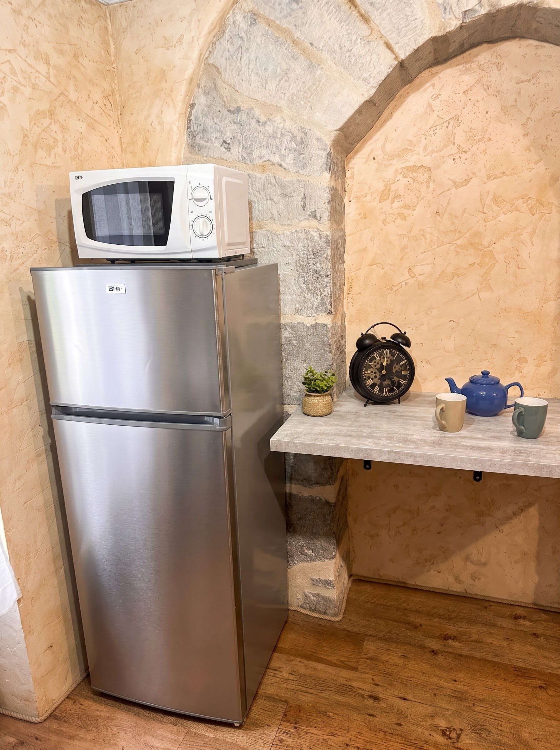 A stainless steel refrigerator with a microwave on top, a small potted plant, a vintage alarm clock, and a blue teapot on a shelf.