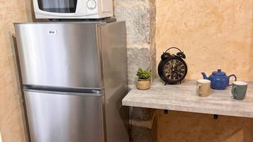 A stainless steel refrigerator with a microwave on top, a small potted plant, a vintage alarm clock, and a blue teapot on a shelf.