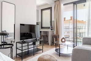 A modern living room with a flat-screen TV, a coffee table, a sofa, and a balcony with a view of buildings. - Yamas Urban Living Olympia Suites Top Floor (Lemesos)