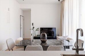 A modern dining area with a marble table, chairs, a vase, a television, and a large window with curtains. - Yamas Urban Living Sequoia 2 BR 2 Bath (Lemesos)