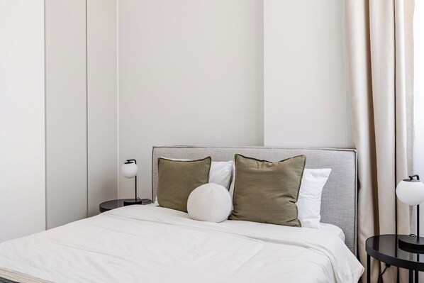 A neatly made bed with a grey headboard, two bedside tables with lamps, and a large window with curtains. - Yamas Urban Living Sequoia 2 BR 2 Bath (Lemesos)