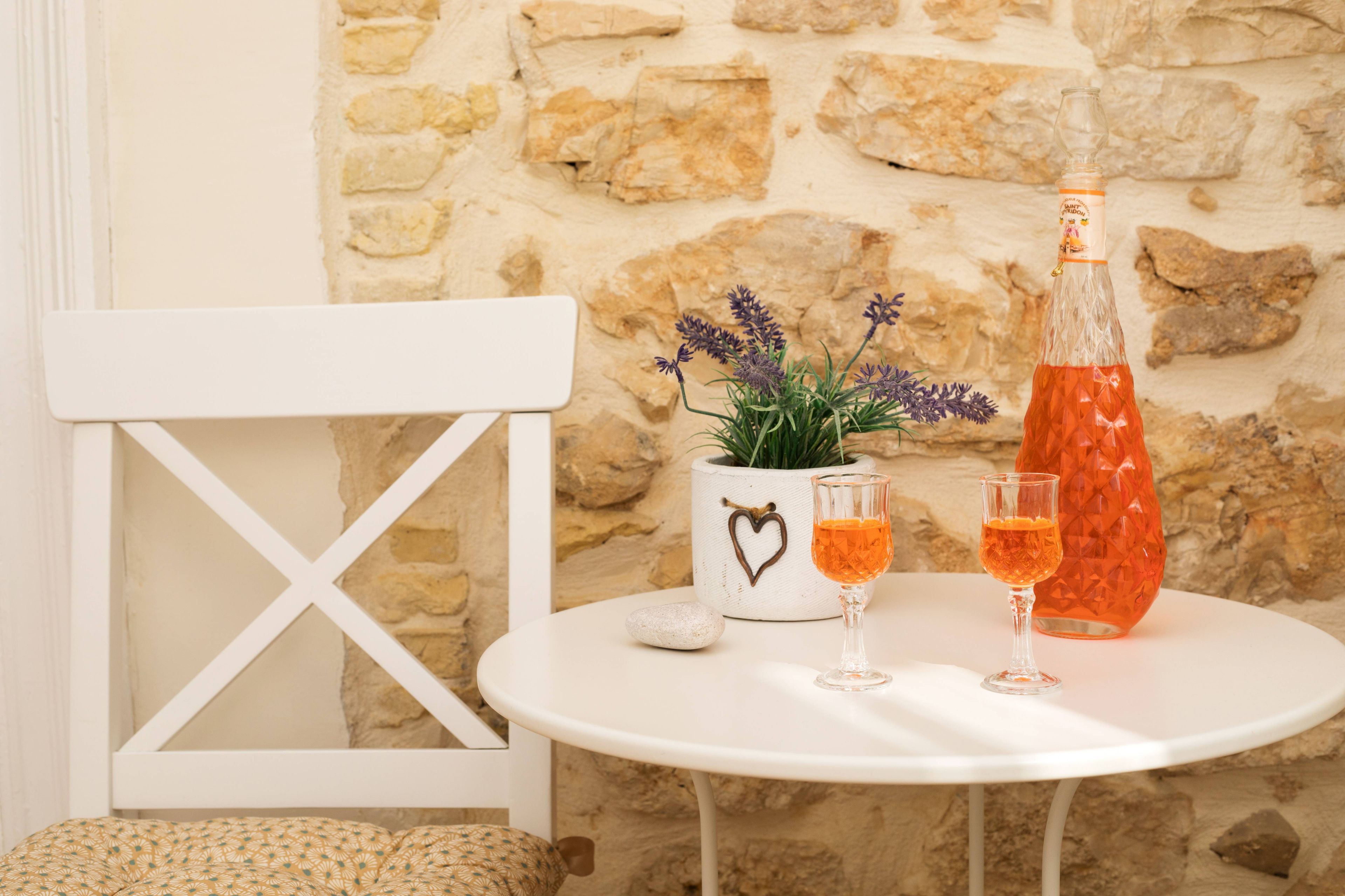 A white table with a stone wall background, a white chair, a potted plant, a bottle, and two glasses.