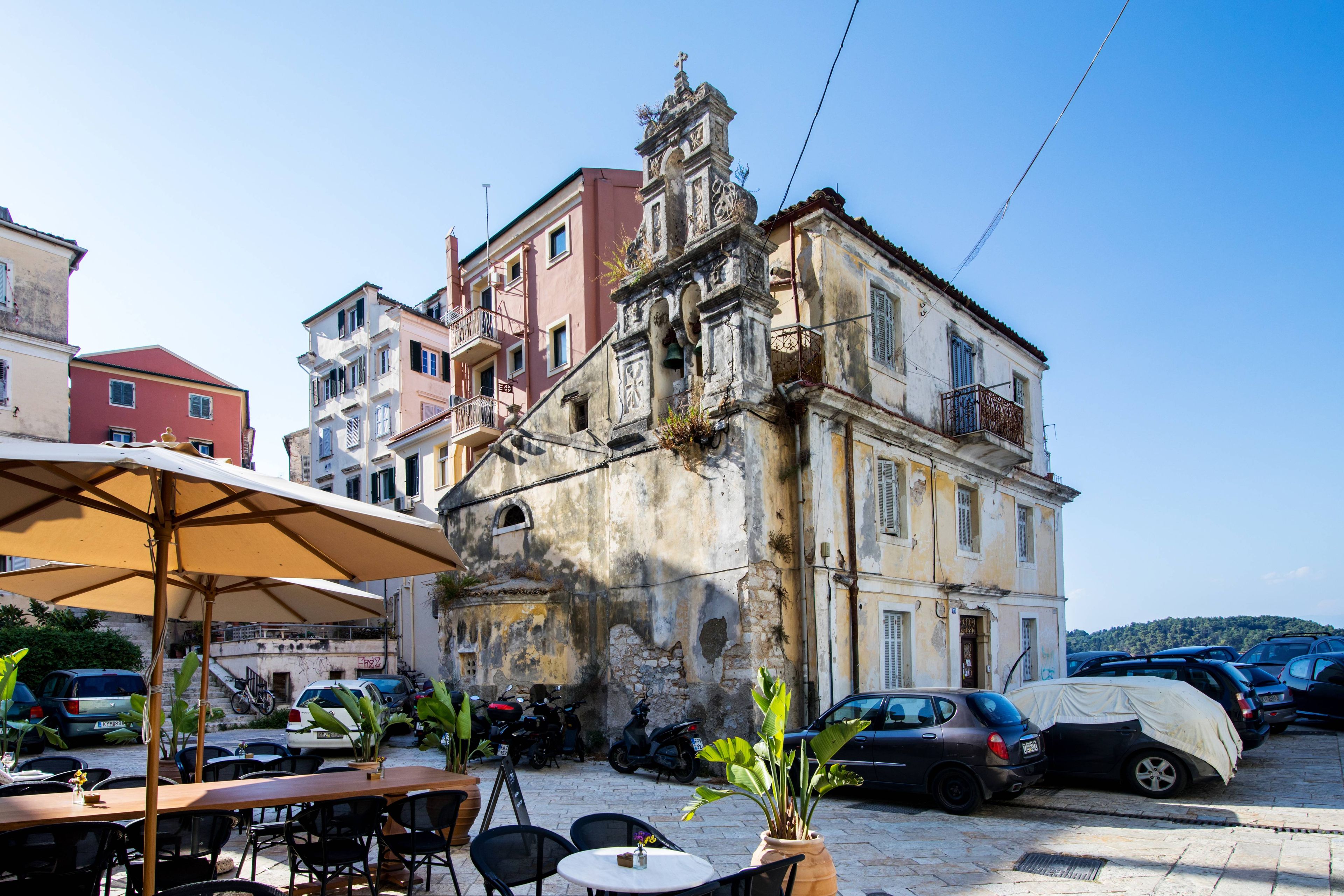 Un café al aire libre con mesas y sillas, un edificio con balcones y coches aparcados.