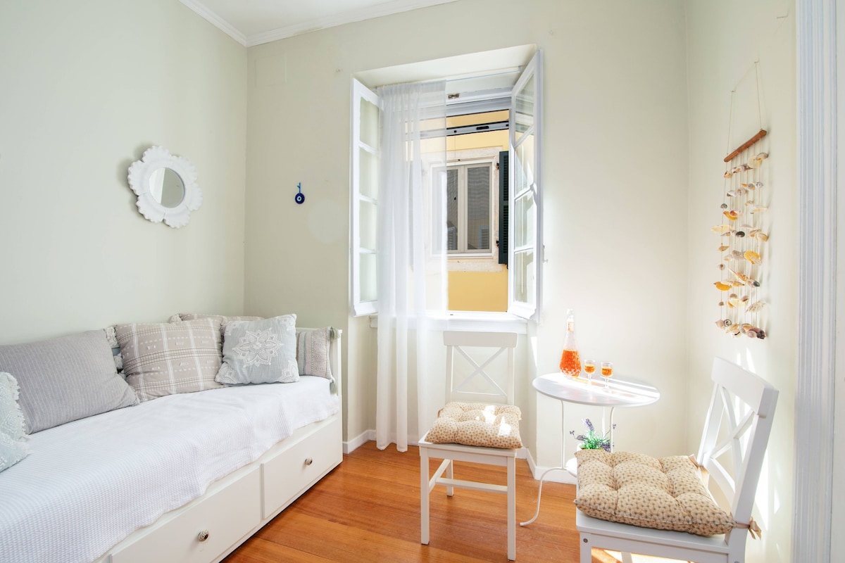 Ein Schlafzimmer mit einem Bett, einem kleinen Tisch mit Getränken, einem Sessel und einem Fenster mit Vorhängen.