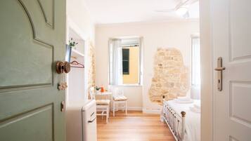 Un pasillo estrecho que conduce a un dormitorio con pared de piedra, una pequeña mesa con una silla y una cama con ropa blanca.