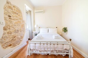A bedroom with a stone wall, a bed with white linens, a bedside table, a lamp, and a potted plant. - Venetian Window Apartments CorfuEscapes (Corfu)
