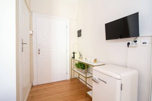 Ein gemütlicher Raum mit weißer Tür, einer kleinen Küchenzeile mit Kühlschrank, einem an der Wand montierten Fernseher und Holzboden.