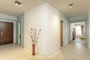Een ruime gang met een grote witte muur, een houten deur en een vaas met takken.