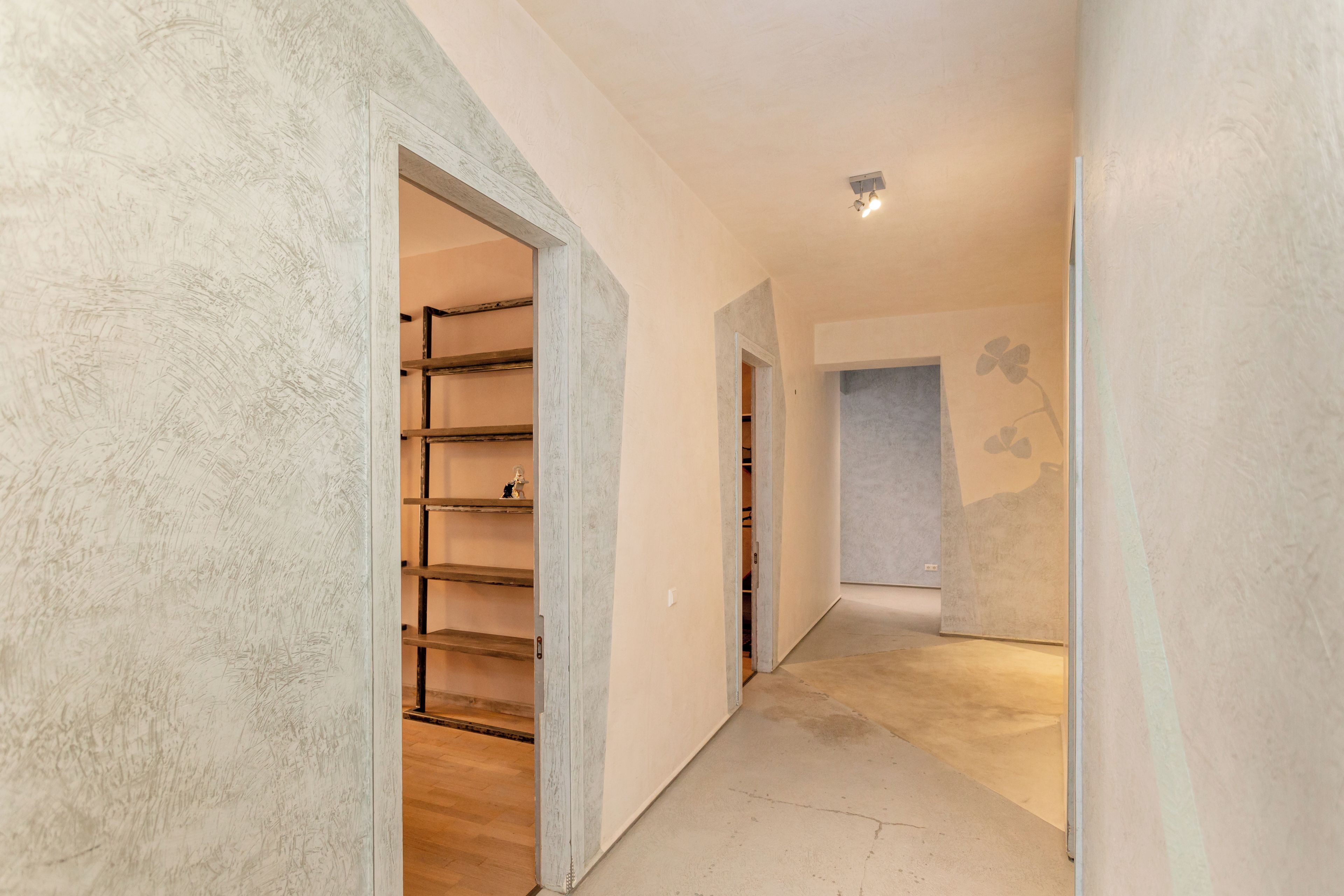 Un couloir doté d’un plancher en bois, d’une étagère en métal et d’une porte donnant sur une pièce dont le mur est peint dans une couleur claire.
