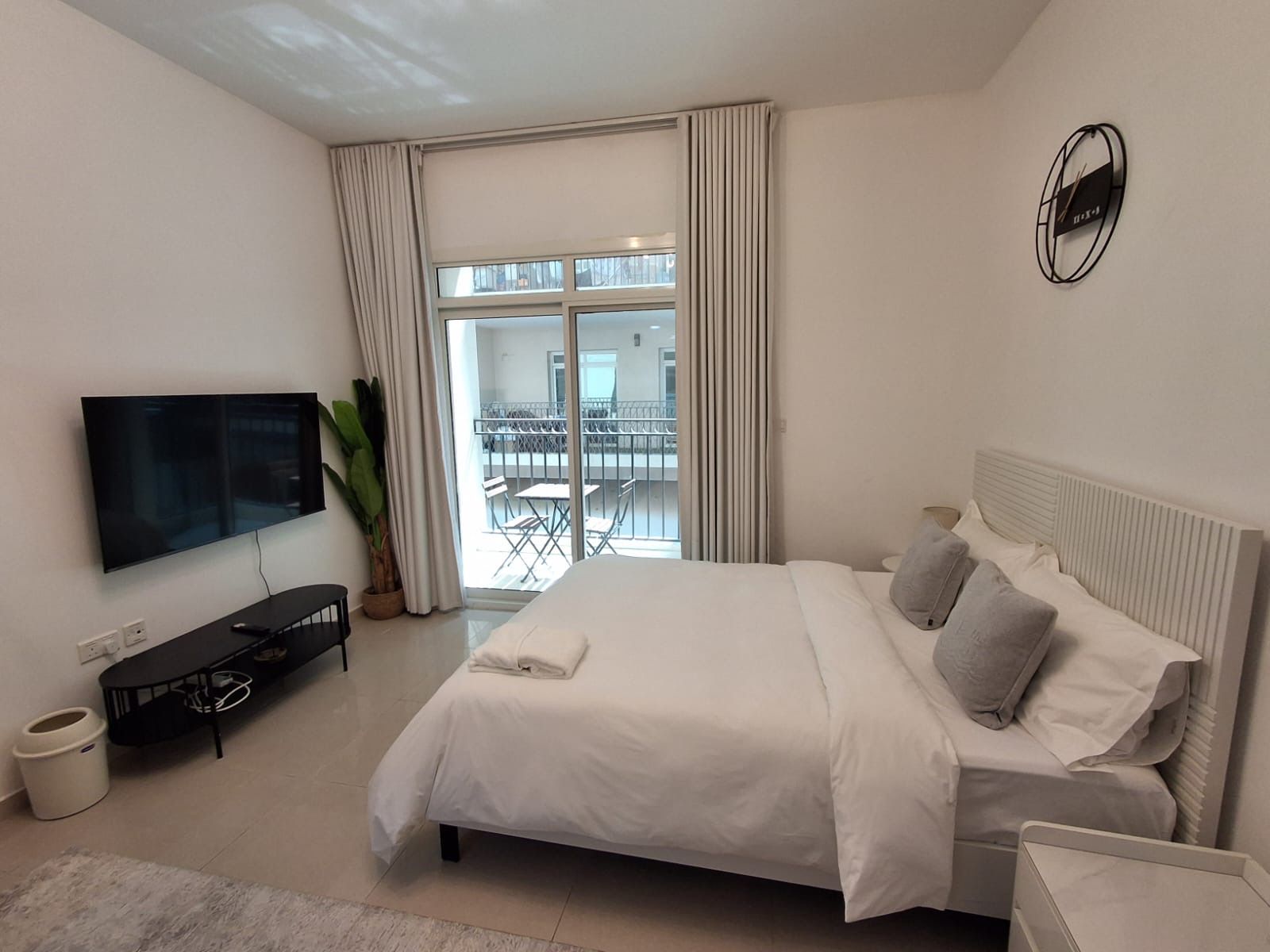 A modern hotel room with a large bed, a flat-screen TV, a balcony with a table and chairs, and a view of the city.