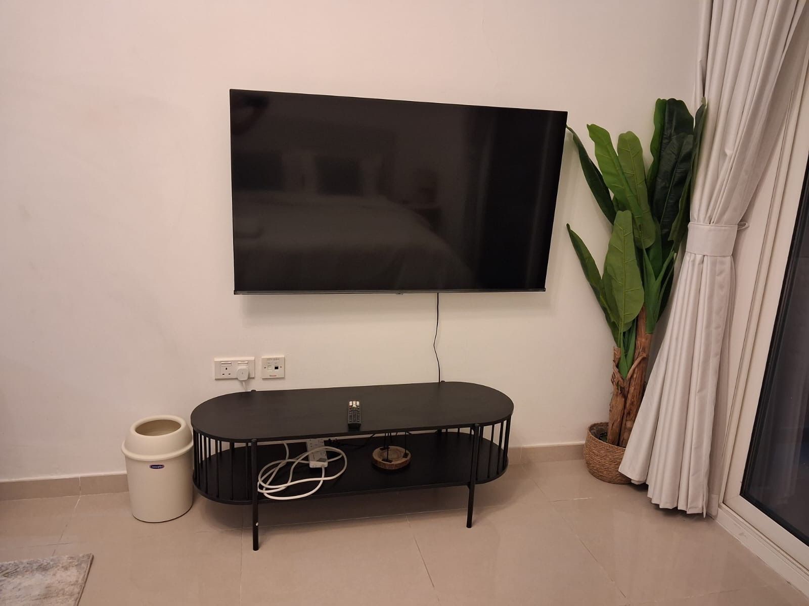 A flat-screen TV mounted on a wall, a black entertainment unit with a remote, a white trash bin, and a potted plant.