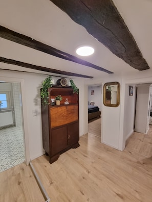 Un couloir doté d’un plancher en bois, une armoire en bois sombre ornée d’objets décoratifs, un miroir accroché au mur et une porte donnant sur une autre pièce.