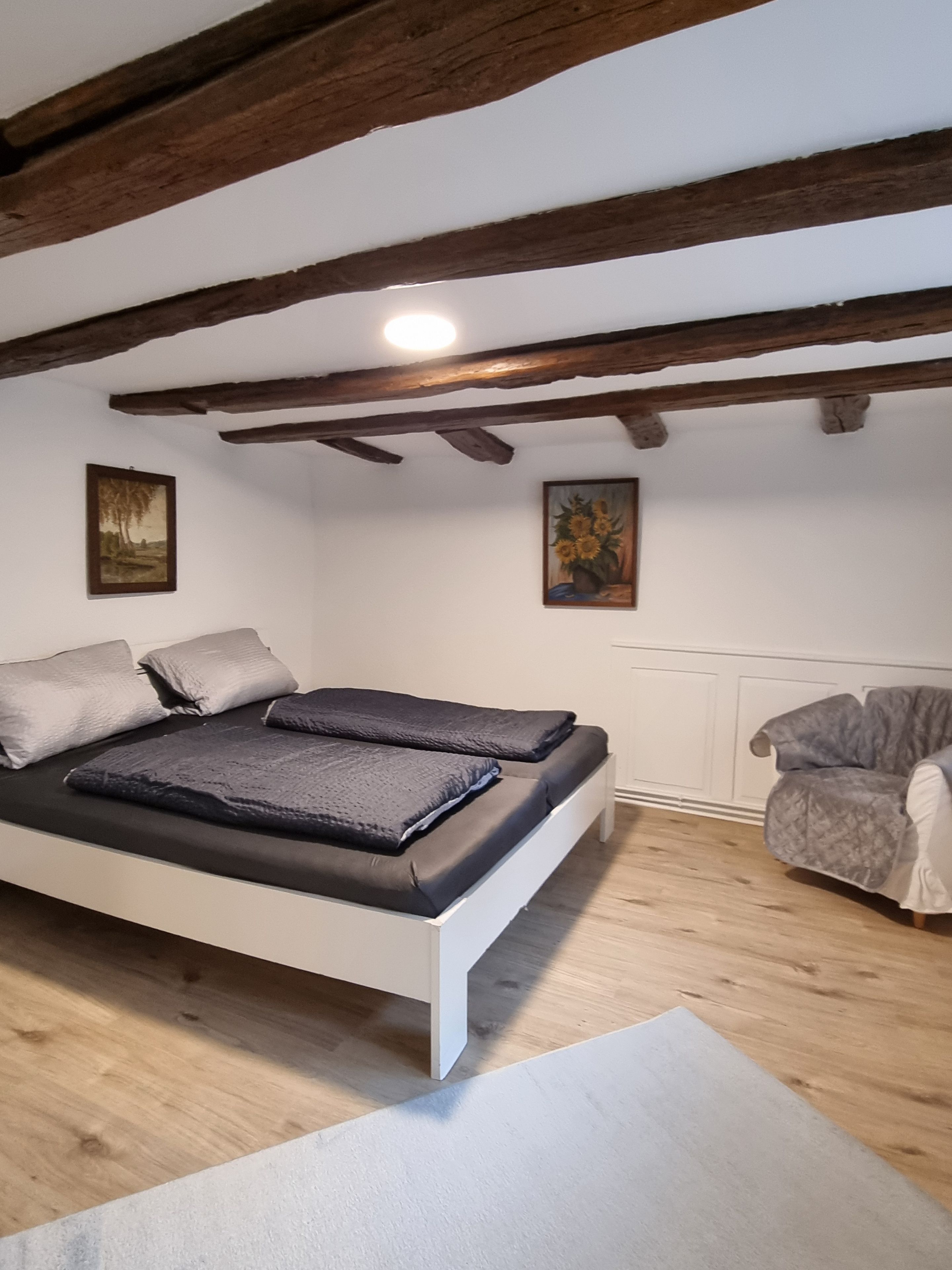 Un dormitorio con cama, una silla, vigas de madera y dos cuadros en la pared.