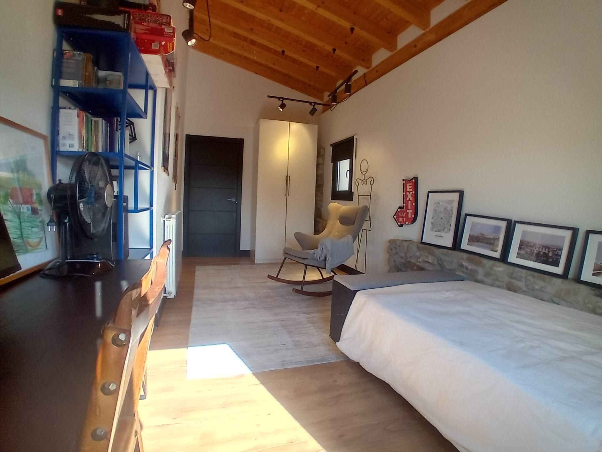 Une chambre à coucher comprenant un lit, un fauteuil à bascule, une armoire et un bureau avec un ventilateur.