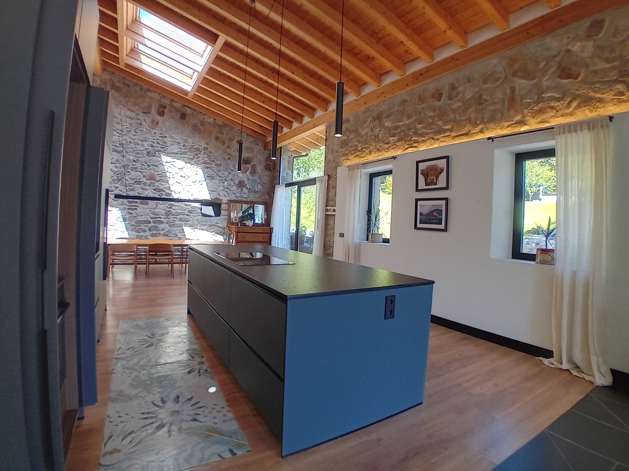 Una cocina moderna con una isla central, paredes de piedra, vigas de madera en el techo y ventanas amplias.