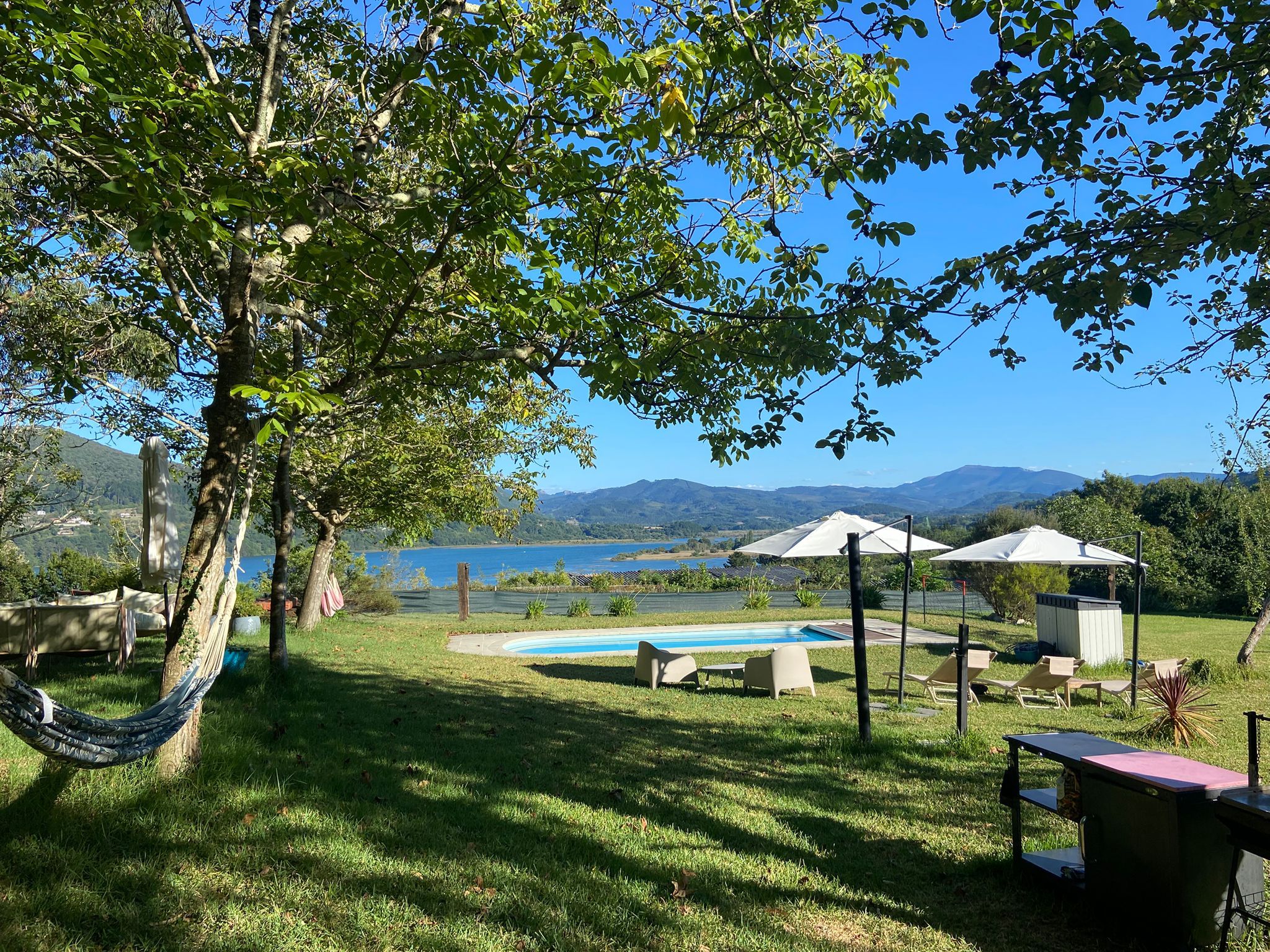 A pool area with lounge chairs and umbrellas, surrounded by grass and trees, with a view of mountains and a body of water in the background.
