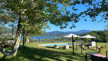 A pool area with lounge chairs and umbrellas, surrounded by grass and trees, with a view of mountains and a body of water in the background.