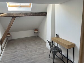 Une pièce avec un plafond en pente, une lucarne, une poutre en bois, une chaise grise, une petite table et une plante en pot.