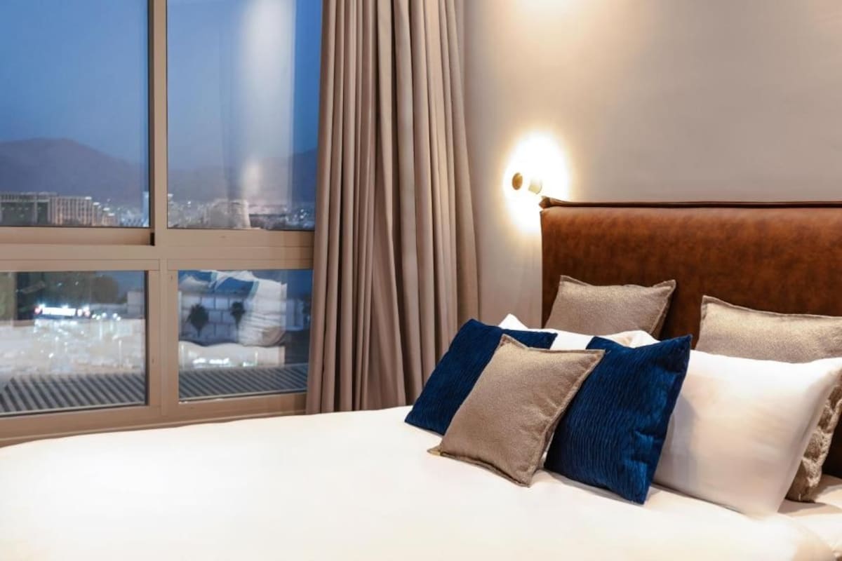A hotel room with a large bed, a view of a cityscape through a window, and a wall-mounted light fixture.