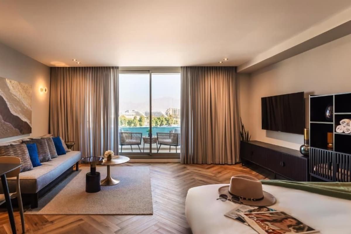 A modern hotel room with a sofa, dining table, and a view of a pool and cityscape through large windows.