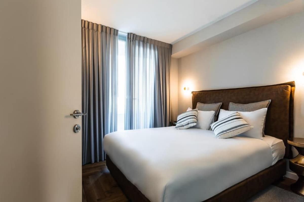 A hotel room with a large bed, curtains, a nightstand, and a wall-mounted light.