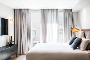 A modern hotel room with a large bed, a flat-screen TV, a bedside lamp, and a view of the city through floor-to-ceiling windows. - Sphera Eilat (Eilat)