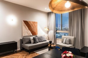 Una sala de estar moderna con un sofá, una mesa de comedor con un florero y vistas a la ciudad a través de grandes ventanales.