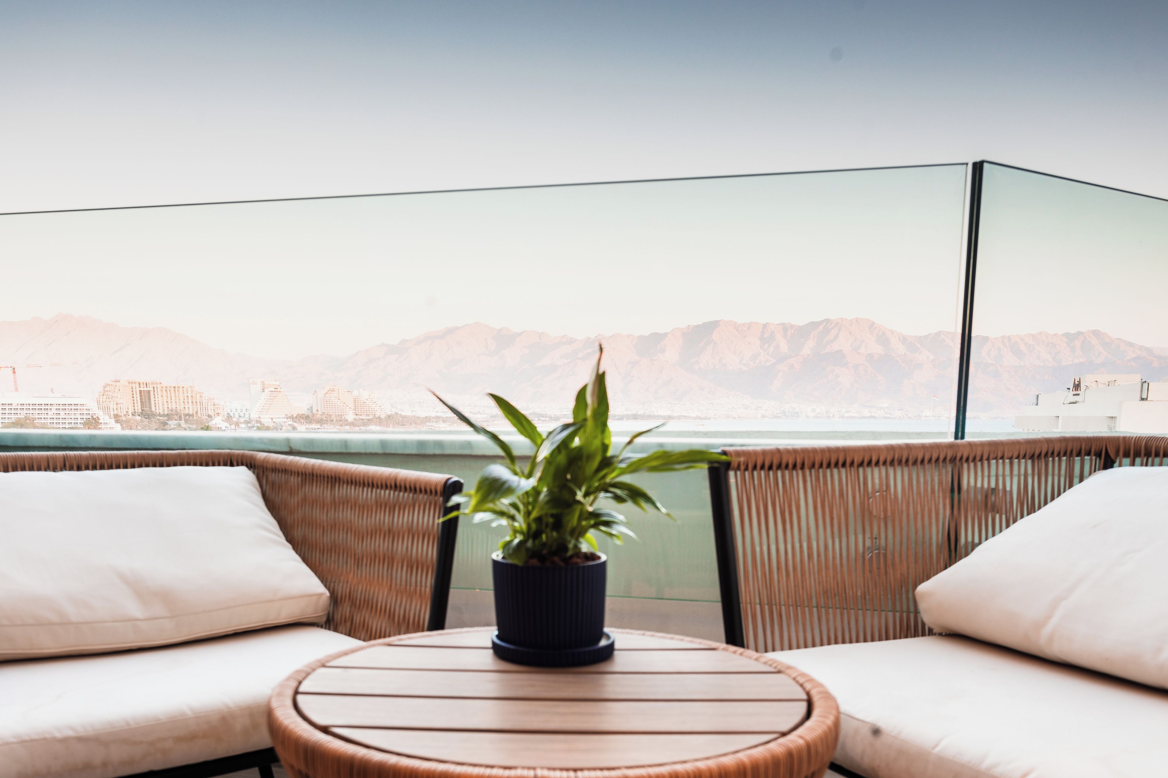 Un balcón con una mesa redonda de madera y dos sillas blancas con cojines, una planta en maceta y vistas a montañas.