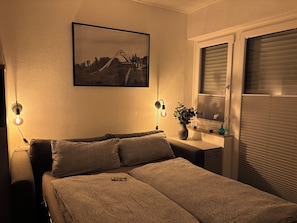 A bedroom with a bed, two bedside lamps, a window with blinds, a vase with a plant, and a framed picture on the wall. - New Listing (Winterberg)